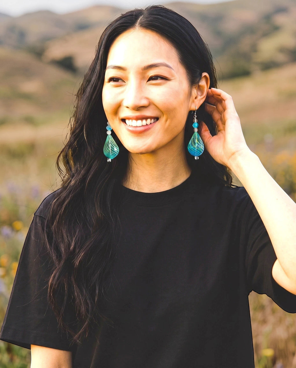Heartfelt Creations handcrafted teal teardrop glass earrings with lime-green swirls worn by a smiling woman outdoors.