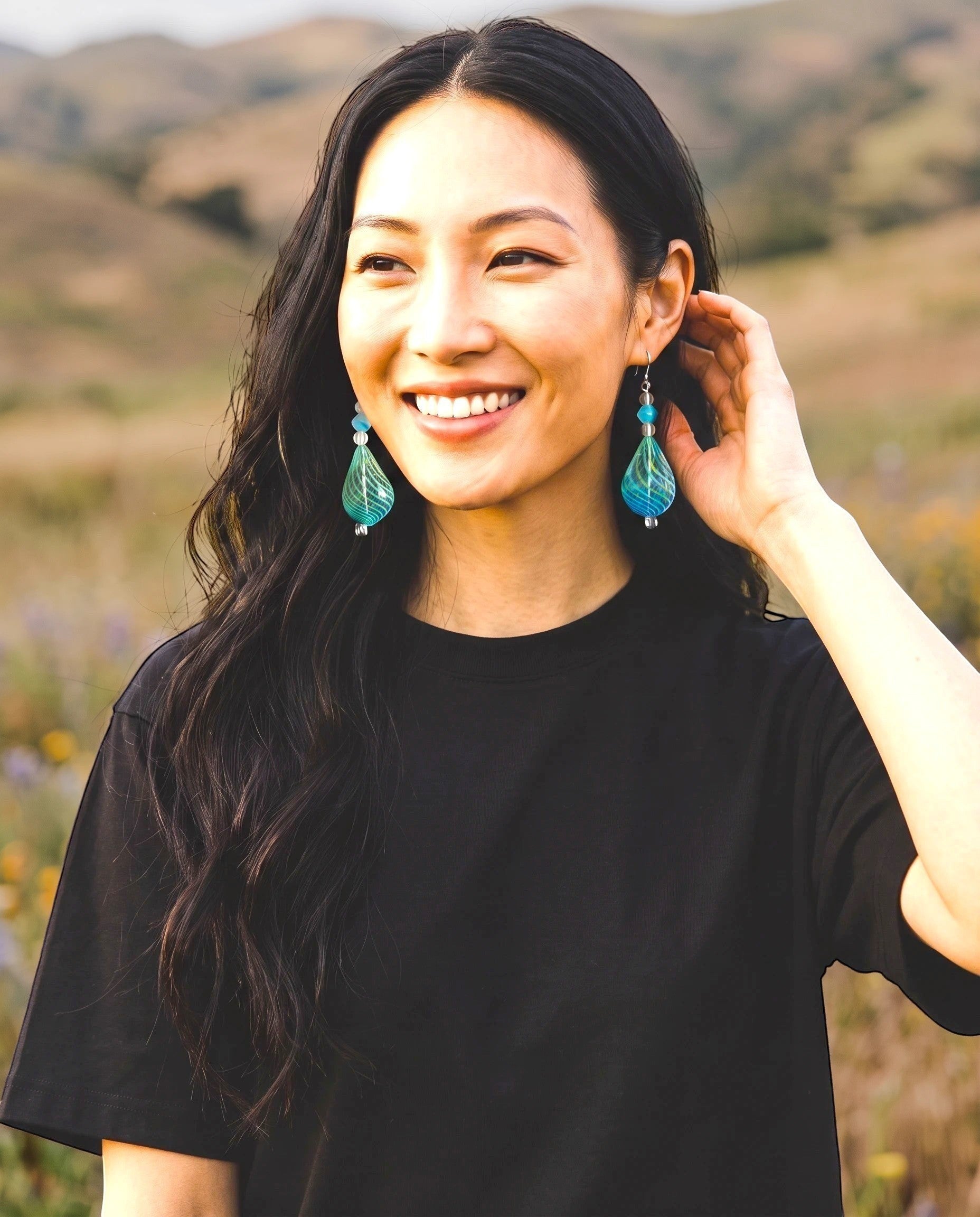 Heartfelt Creations handcrafted teal teardrop glass earrings with lime-green swirls worn by a smiling woman outdoors.