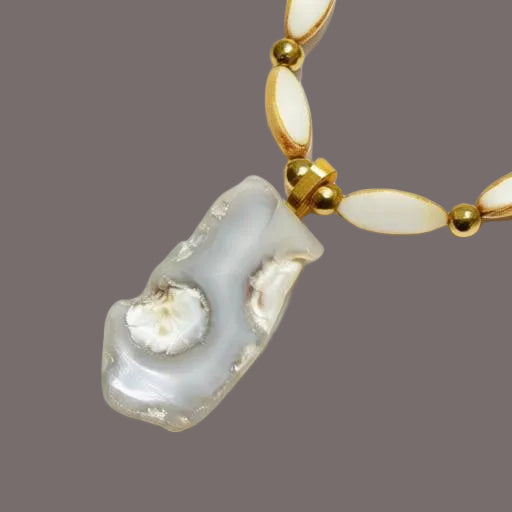 A close-up of a necklace featuring a large, irregular polished agate pendant with natural matrix edges and a white inlay, accented by gold beads and oval white inlay spacers on a white background.