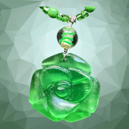 A green translucent carved pendant shaped like a rose, hanging from a matching green beaded necklace with a faceted green bead and a clear bead containing green swirls, set against a gray geometric background.