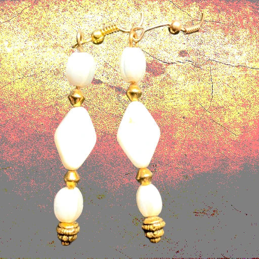 A pair of dangling earrings with large white oval beads, smaller gold-tone spacer beads, and textured gold-tone end caps, shown against a mottled gold, red, and gray background.