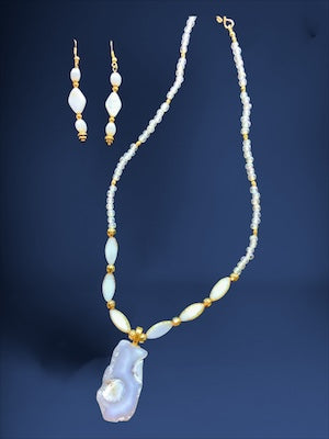 A beaded necklace and matching drop earrings displayed on a dark blue background. The jewelry features white beads, gold-toned metal spacers and hooks, and a large irregular white pearl pendant.