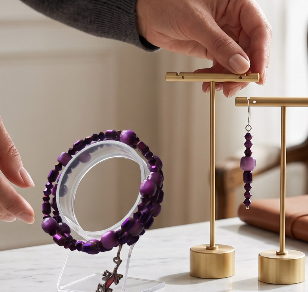 A person's hands display a pair of purple beaded drop earrings and a matching purple beaded bracelet on minimalist gold jewelry stands, set on a white marble tabletop in a bright, modern room.