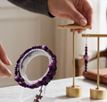 A person's hands display a pair of purple beaded drop earrings and a matching purple beaded bracelet on minimalist gold jewelry stands, set on a white marble tabletop in a bright, modern room.