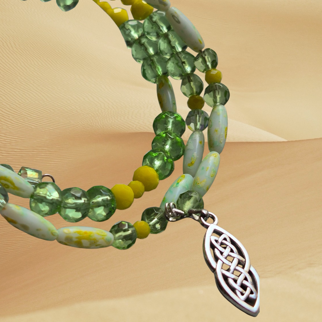A close-up of a beaded strand jewelry piece with green faceted beads, pale green marbled beads, small yellow pom-pom beads, and a silver Celtic knot leaf-shaped charm, set against a sandy beige background.