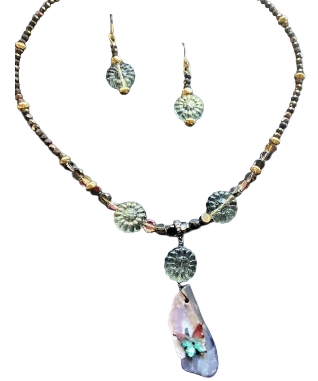 A handcrafted necklace and earring set featuring a delicate beaded chain of small, dark grey and golden beads, with a central pendant made of a polished, iridescent purple and white shell piece adorned with a small, colorful butterfly motif, accompanied by matching earrings with a single, translucent, light green floral-shaped bead