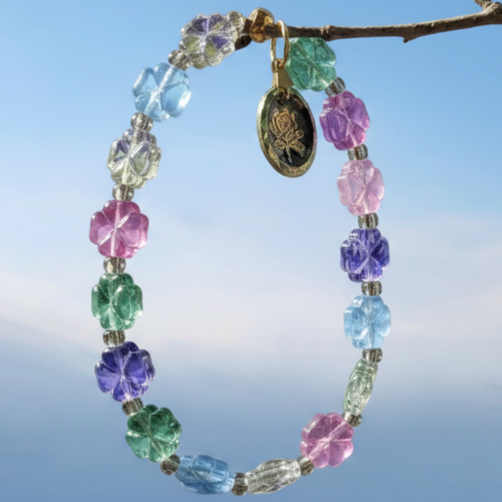 A beaded bracelet with colorful faceted flower-shaped beads in pink, purple, blue, green, and clear, hanging from a thin brown branch against a soft blue sky background. A round gold-toned charm hangs from the top.