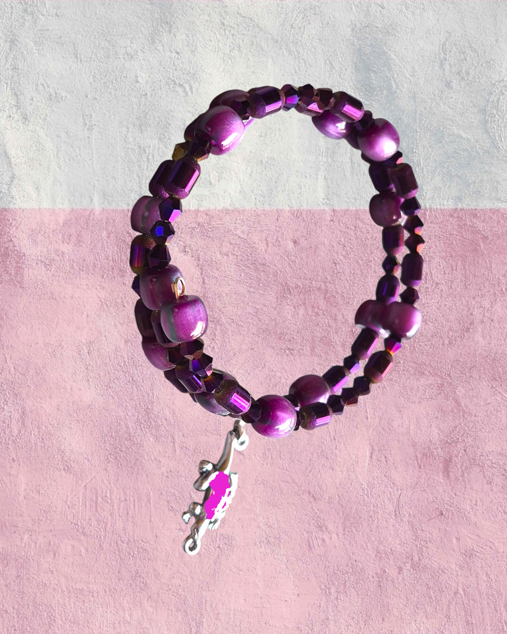 A beaded stretch bracelet featuring purple and magenta beads with a small silver branch charm, displayed against a split white and pink textured background.