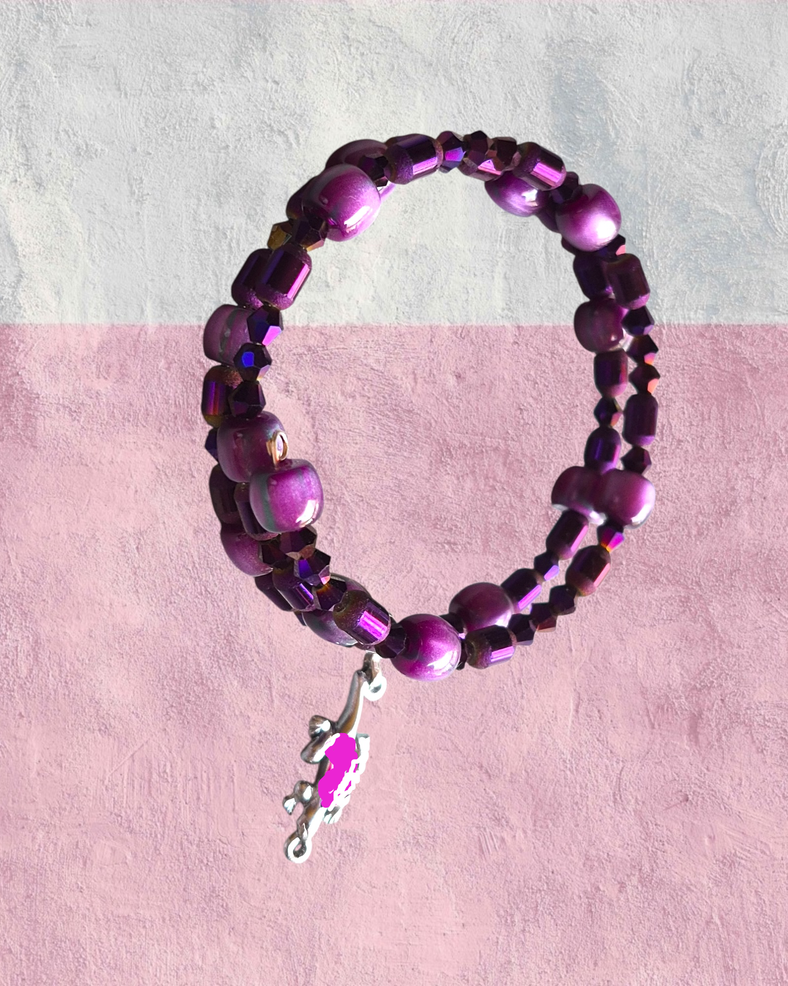 A beaded stretch bracelet featuring purple and magenta beads with a small silver branch charm, displayed against a split white and pink textured background.
