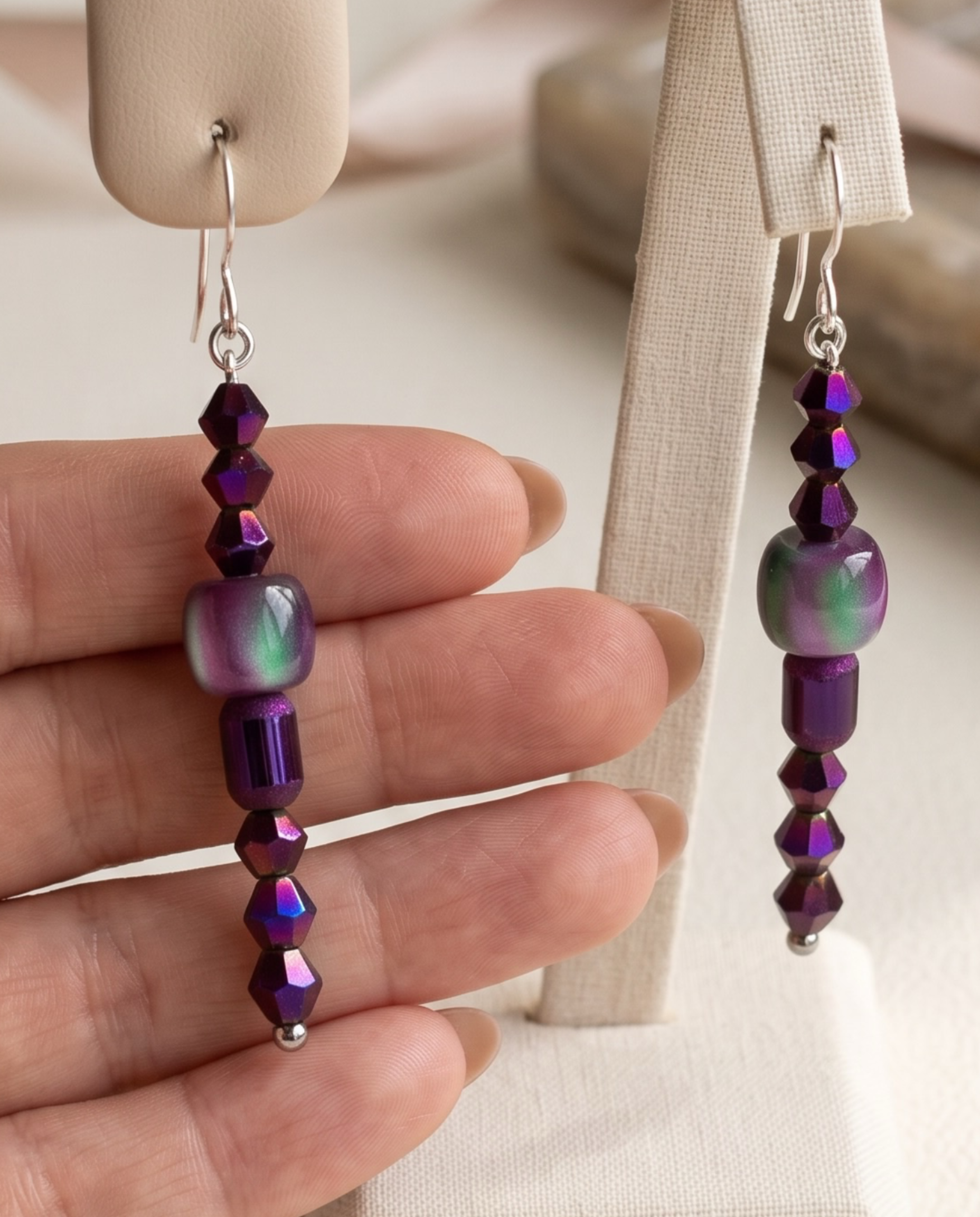 A pair of silver-toned dangle earrings with textured disc and bead details, a knotted rope-like drop, and polished ball ends, shown against a purple studio background.