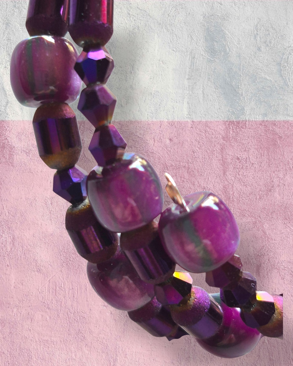 Purple iridescent beaded necklace with large round beads and faceted beads, displayed on a textured pink and white background.