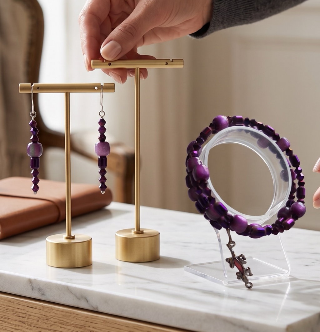 A person's hands display a pair of purple beaded drop earrings and a matching purple beaded bracelet on minimalist gold jewelry stands, set on a white marble tabletop in a bright, modern room.