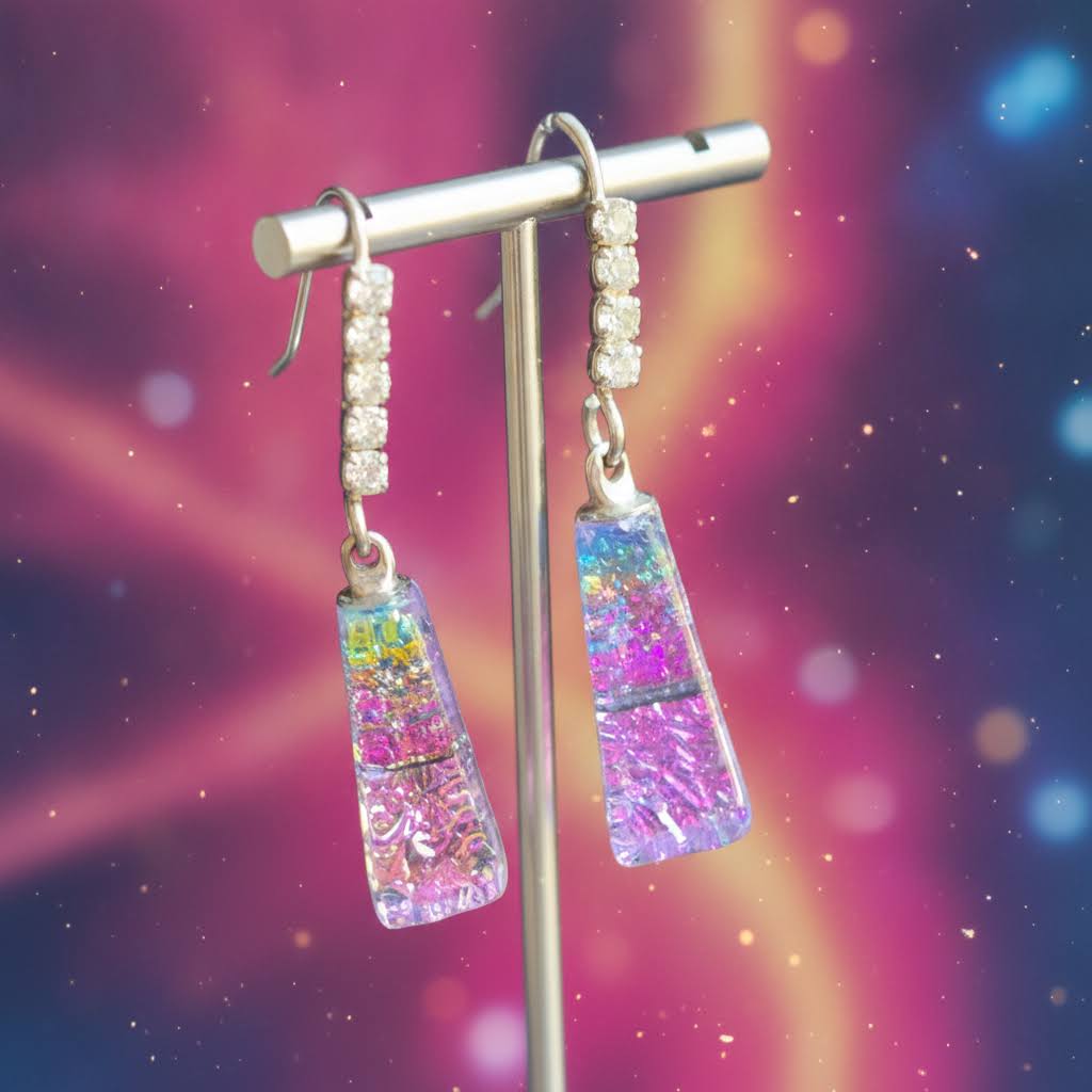 A pair of dangling earrings displayed on a metal stand against a colorful cosmic background. The earrings feature silver-toned metal hooks with small clear stones, and rectangular glass drops filled with pink, purple, blue, and green glitter.