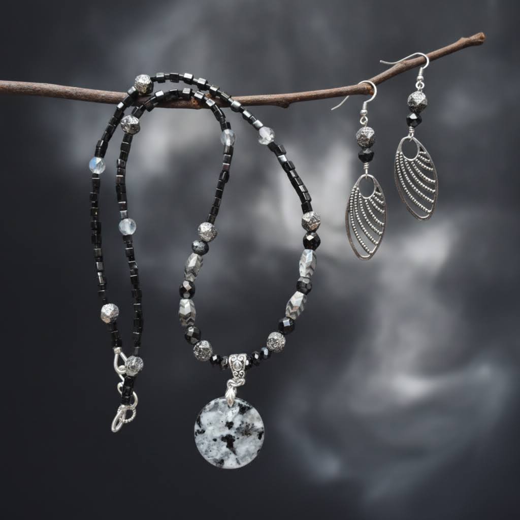 Heartfelt Creations  handmade jewelry set featuring a necklace and matching earrings, crafted from various black, white, and silver beads, with a circular, speckled granite-like stone pendant on the necklace and intricate, black filigree leaf-shaped pendants on the earrings.