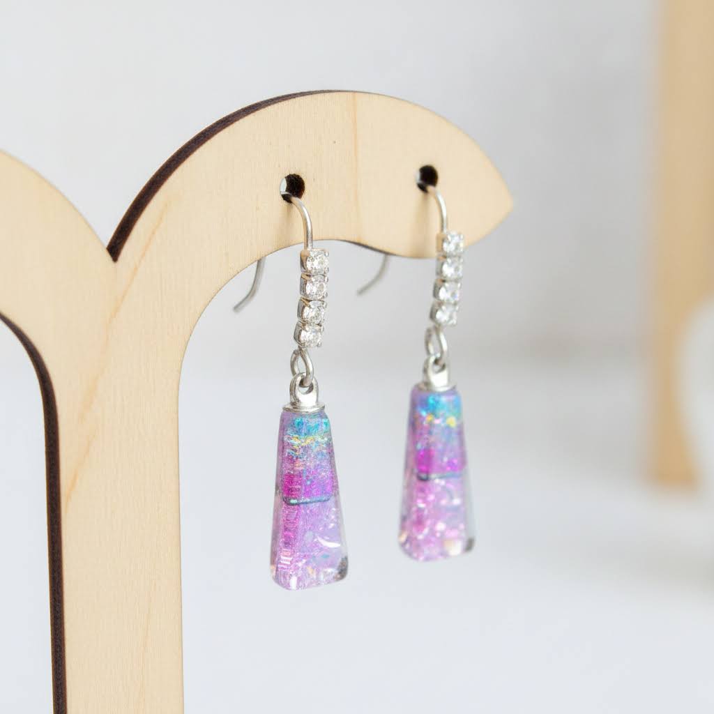 A pair of dangling earrings displayed on a light wood stand. Each earring has a silver fishhook with a small clear rhinestone-studded section and a tapered clear resin drop filled with pink, purple, and iridescent glitter.
