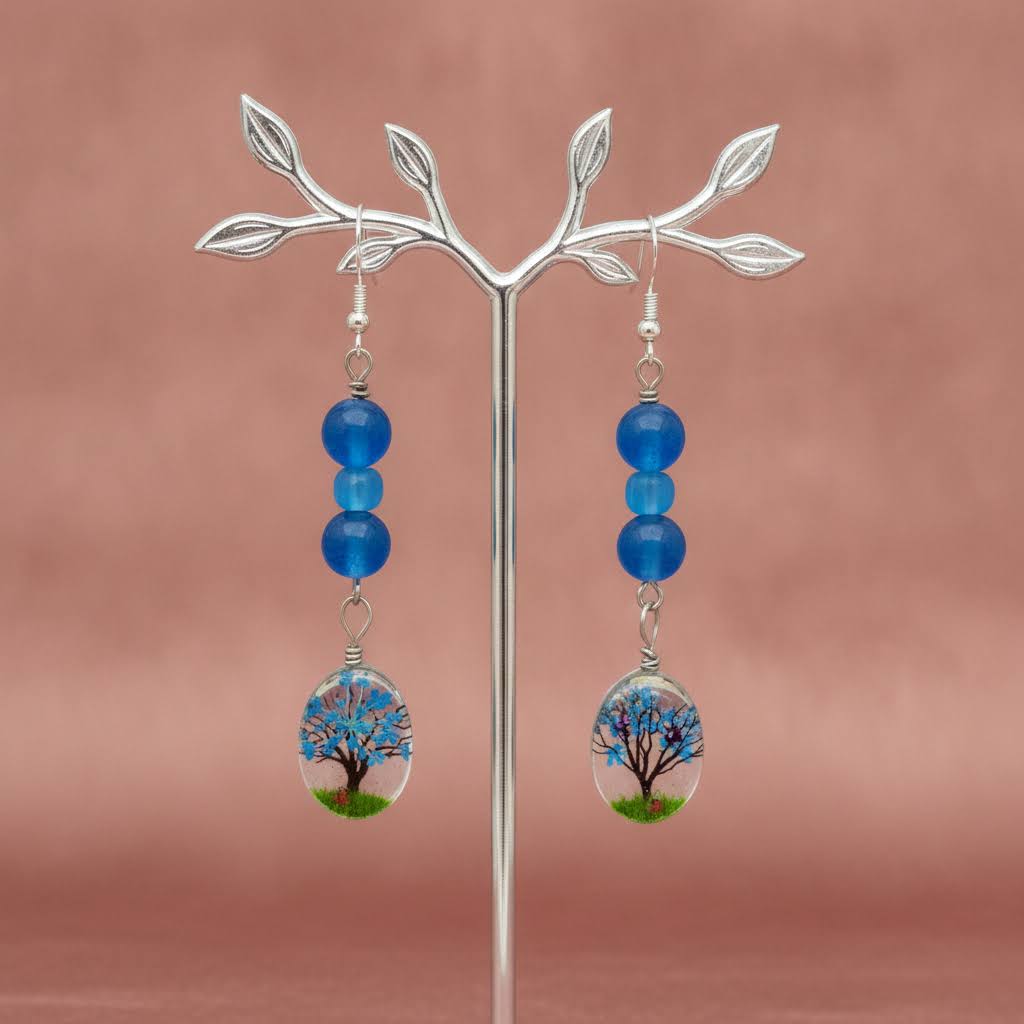 A pair of dangling earrings displayed on a silver branch stand against a dusty pink background. Each earring has three blue beads hanging from a silver hook, with an oval clear pendant containing a miniature tree design with blue branches and green grass.