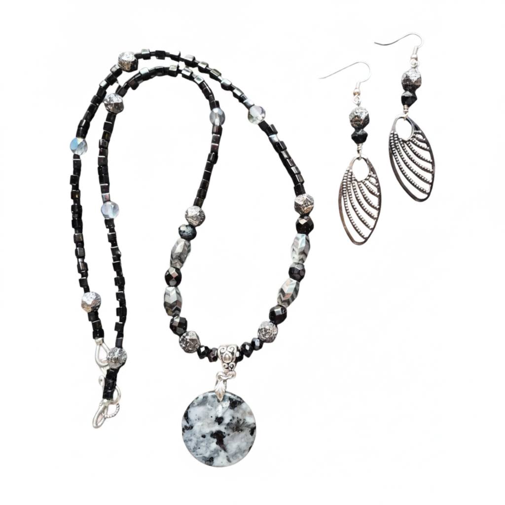 Heartfelt Creations  handmade jewelry set featuring a necklace and matching earrings, crafted from various black, white, and silver beads, with a circular, speckled granite-like stone pendant on the necklace and intricate, black filigree leaf-shaped pendants on the earrings.