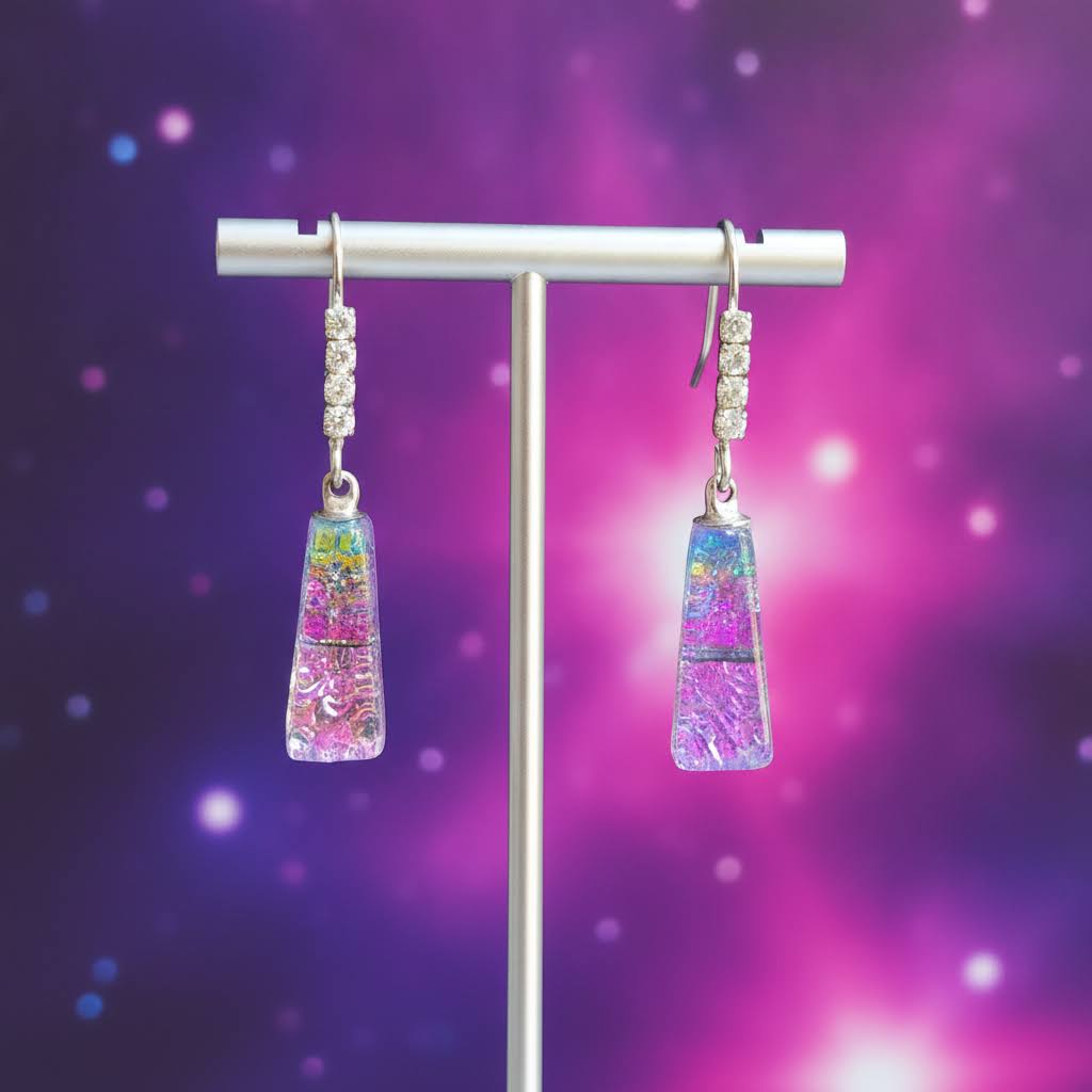 A pair of dangling earrings displayed on a silver stand against a purple and pink bokeh background. The earrings feature rectangular glass pendants filled with colorful swirls of pink, blue, and green, suspended from silver hooks with small clear stones.
