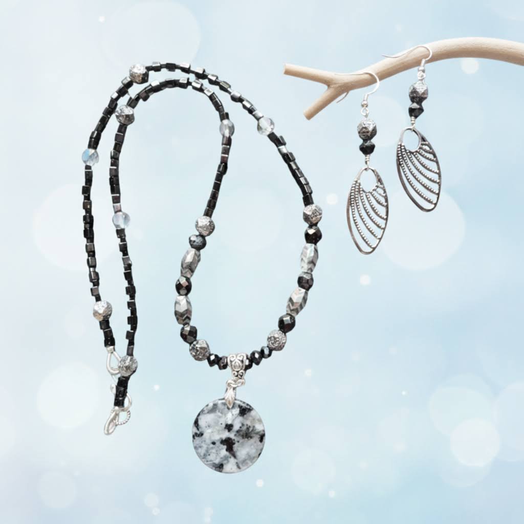 Heartfelt Creations  handmade jewelry set featuring a necklace and matching earrings, crafted from various black, white, and silver beads, with a circular, speckled granite-like agate stone pendant on the necklace and intricate, black filigree leaf-shaped pendants on the earrings.