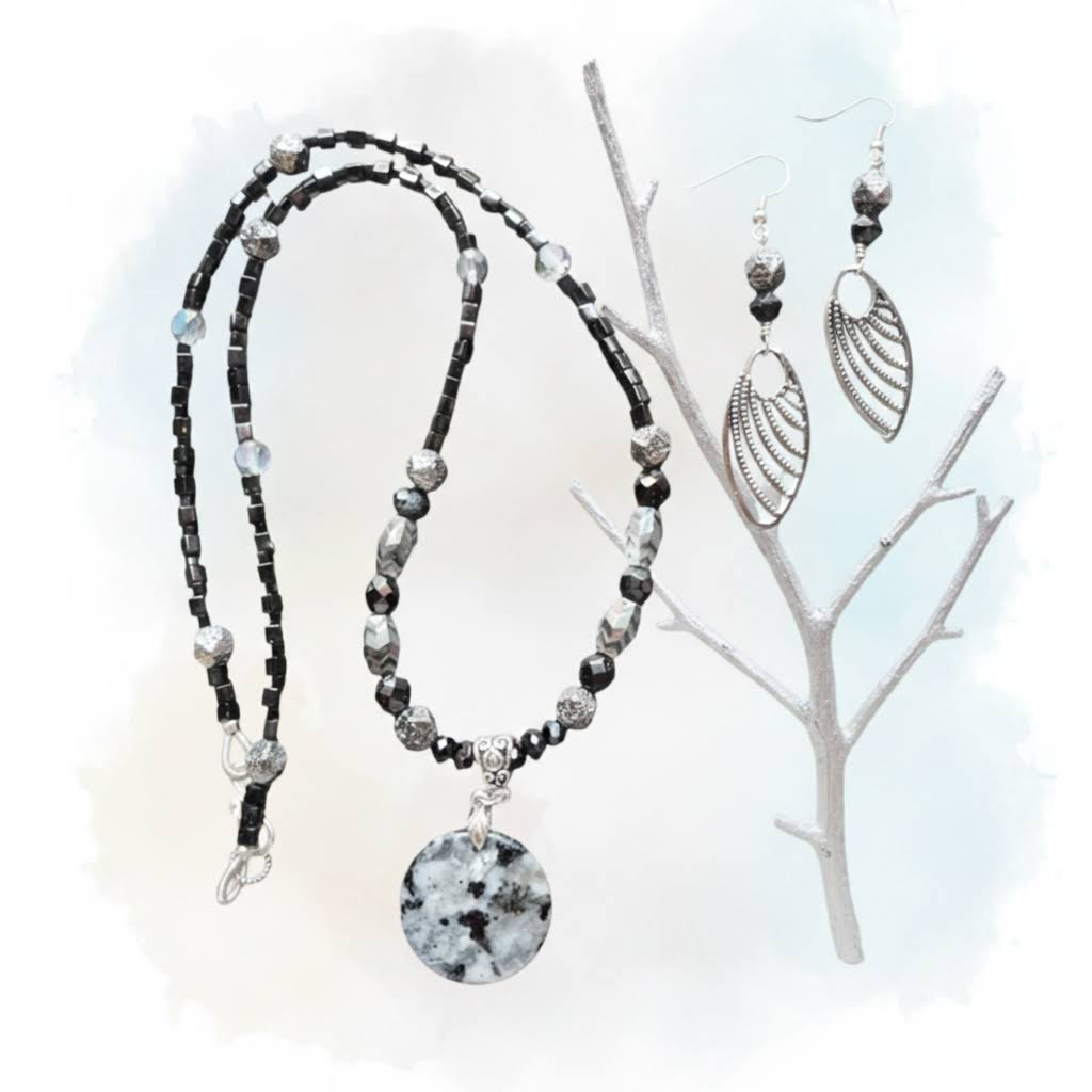 Heartfelt Creations  handmade jewelry set featuring a necklace and matching earrings, crafted from various black, white, and silver beads, with a circular, speckled granite-like agate stone pendant on the necklace and intricate, black filigree leaf-shaped pendants on the earrings.