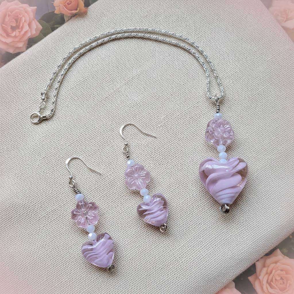 Heartfelt Creations offers a matching set of handcrafted jewelry, including a necklace and a pair of earrings, featuring silver-toned chains and hooks, with each piece adorned with a pale pink, swirled lampwork  glass heart bead and a delicate, clear glass flower bead.