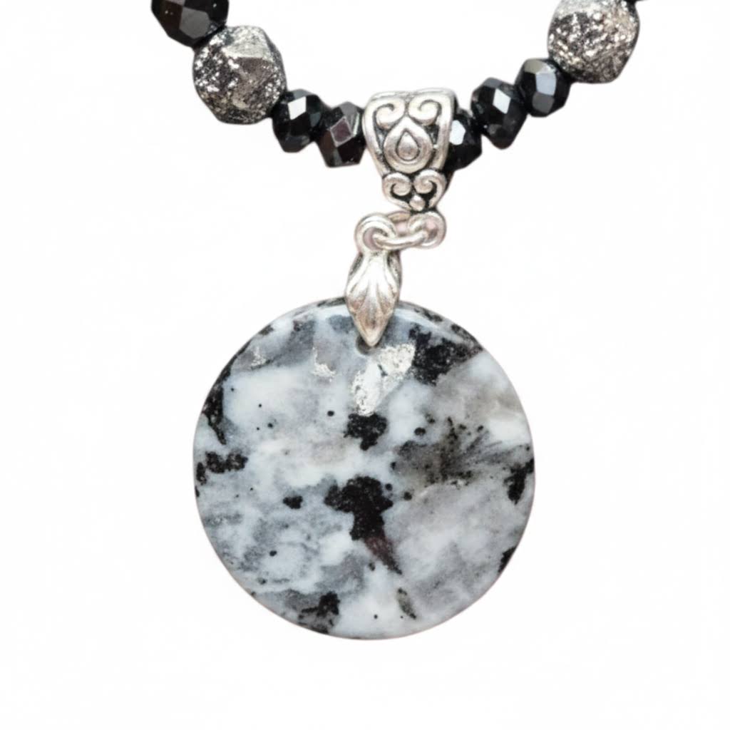 A close-up of a beaded necklace featuring a round white and black marbled agate stone pendant, suspended from a silver-toned decorative clasp and black faceted beads.
