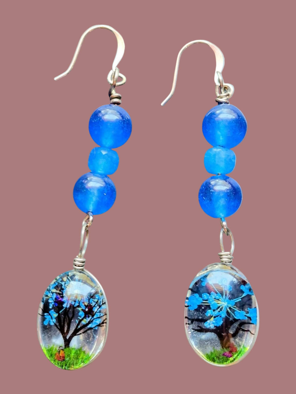 A pair of handmade dangle earrings featuring three blue beads and a clear oval resin charm containing a painted tree and grass scene, set against a solid dusty rose background.