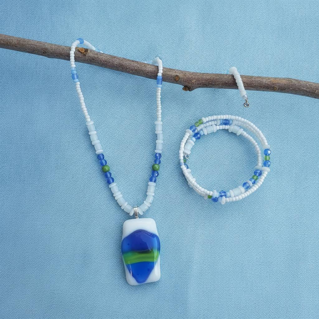 Heartfelt Creations' handmade jewelry set featuring a necklace and a multi-strand bracelet, both crafted with a mix of small, round, white, blue, and green beads, with the necklace featuring a prominent, rectangular, fused glass pendant with a vibrant blue, green, and white abstract design.