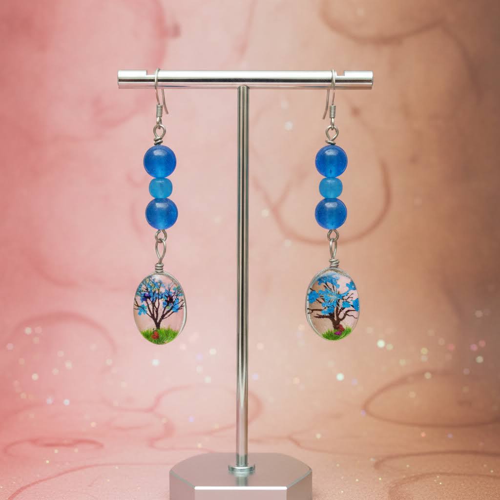 A pair of dangling earrings displayed on a silver stand against a soft pink glittery background. Each earring has three blue beads above an oval clear pendant featuring a painted tree with blue blossoms and green grass.