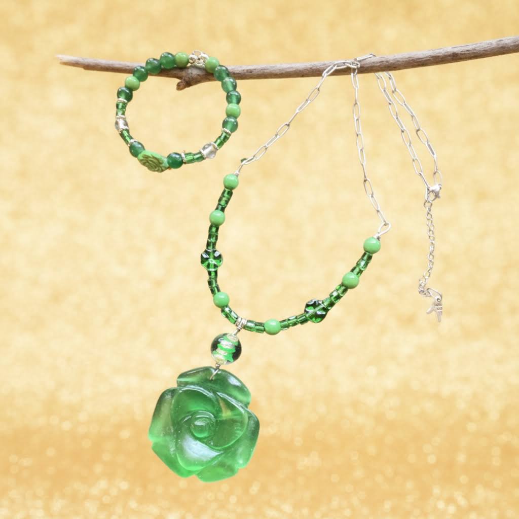 Heartfelt Creations' offers this stunning, nature-inspired beaded necklace and bracelet set. Featuring a vibrant palette of lush greens, this set is designed to add a touch of botanical elegance to any outfit. The centerpiece of the necklace is a beautifully detailed, frosted glass floral charm that captures the essence of a blooming rose, complemented by a variety of green beads that offer a rich, multi-textured look. The matching bracelet echoes this design, creating a cohesive and polished aesthetic.