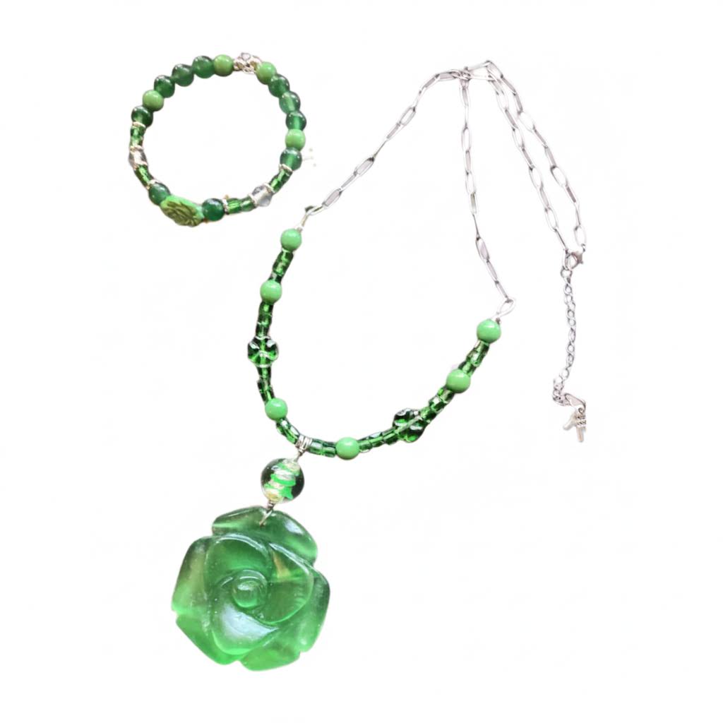 Heartfelt Creations' offers this stunning, nature-inspired beaded necklace and bracelet set. Featuring a vibrant palette of lush greens, this set is designed to add a touch of botanical elegance to any outfit. The centerpiece of the necklace is a beautifully detailed, frosted glass floral charm that captures the essence of a blooming rose, complemented by a variety of green beads that offer a rich, multi-textured look. The matching bracelet echoes this design, creating a cohesive and polished aesthetic.
