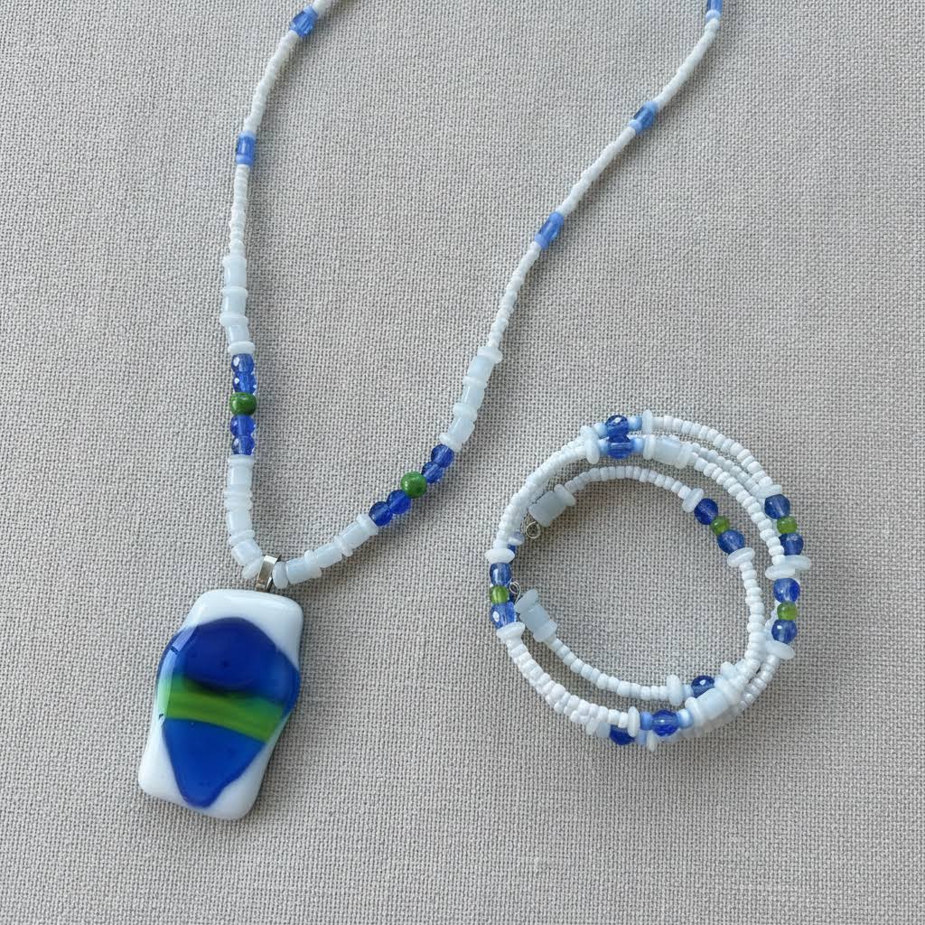 Heartfelt Creations' handmade jewelry set featuring a necklace and a multi-strand bracelet, both crafted with a mix of small, round, white, blue, and green beads, with the necklace featuring a prominent, rectangular, fused glass pendant with a vibrant blue, green, and white abstract design.