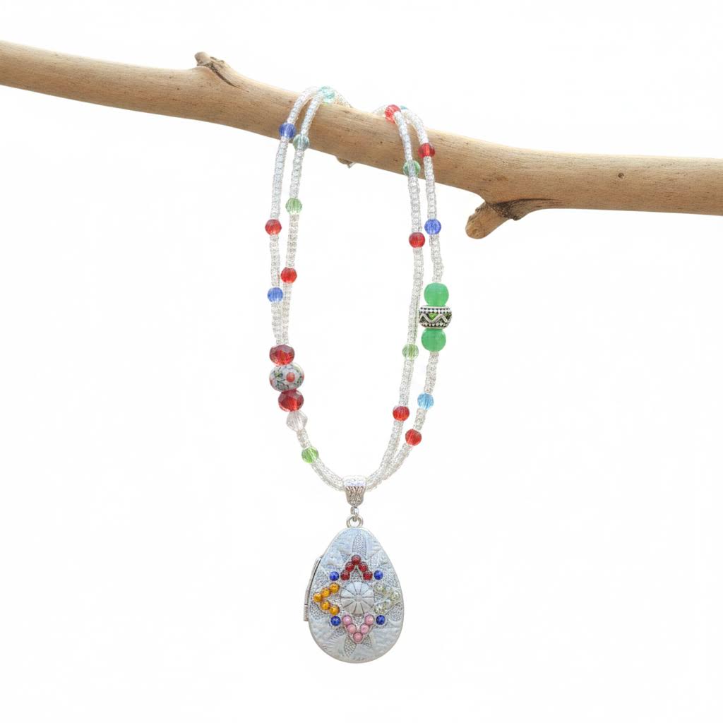 A beaded necklace with white pearl-like strands, colorful red, blue, green, and silver beads, and a large teardrop pendant featuring a colorful floral mandala design, hanging from a natural branch against a white background.