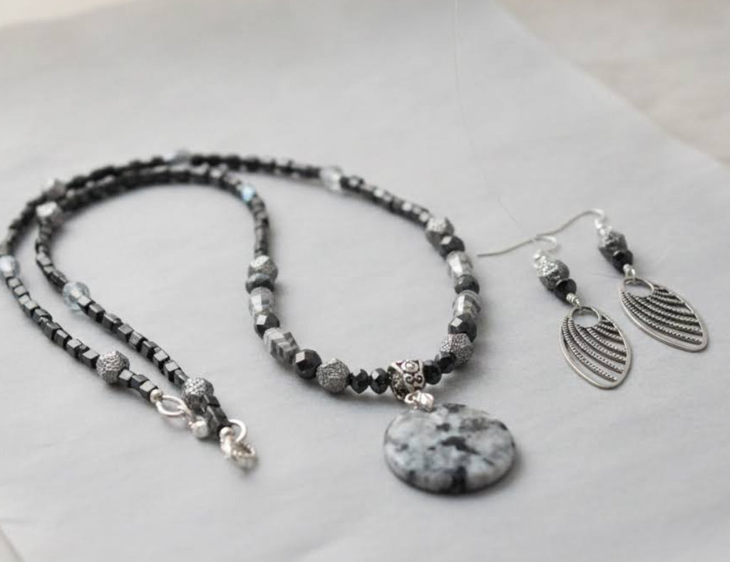 Heartfelt Creations  handmade jewelry set featuring a necklace and matching earrings, crafted from various black, white, and silver beads, with a circular, speckled granite-like agate stone pendant on the necklace and intricate, black filigree leaf-shaped pendants on the earrings.