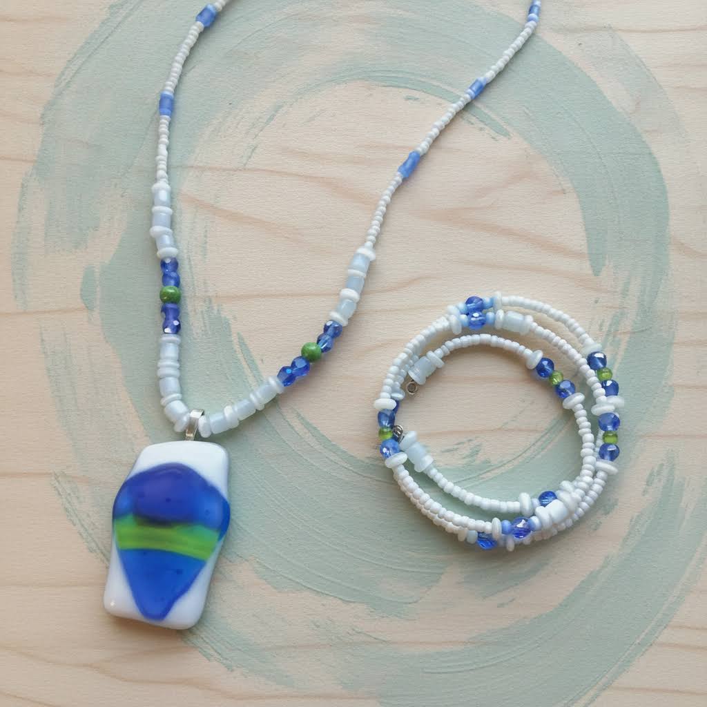 Heartfelt Creations' handmade jewelry set featuring a necklace and a multi-strand bracelet, both crafted with a mix of small, round, white, blue, and green beads, with the necklace featuring a prominent, rectangular, fused glass pendant with a vibrant blue, green, and white abstract design.