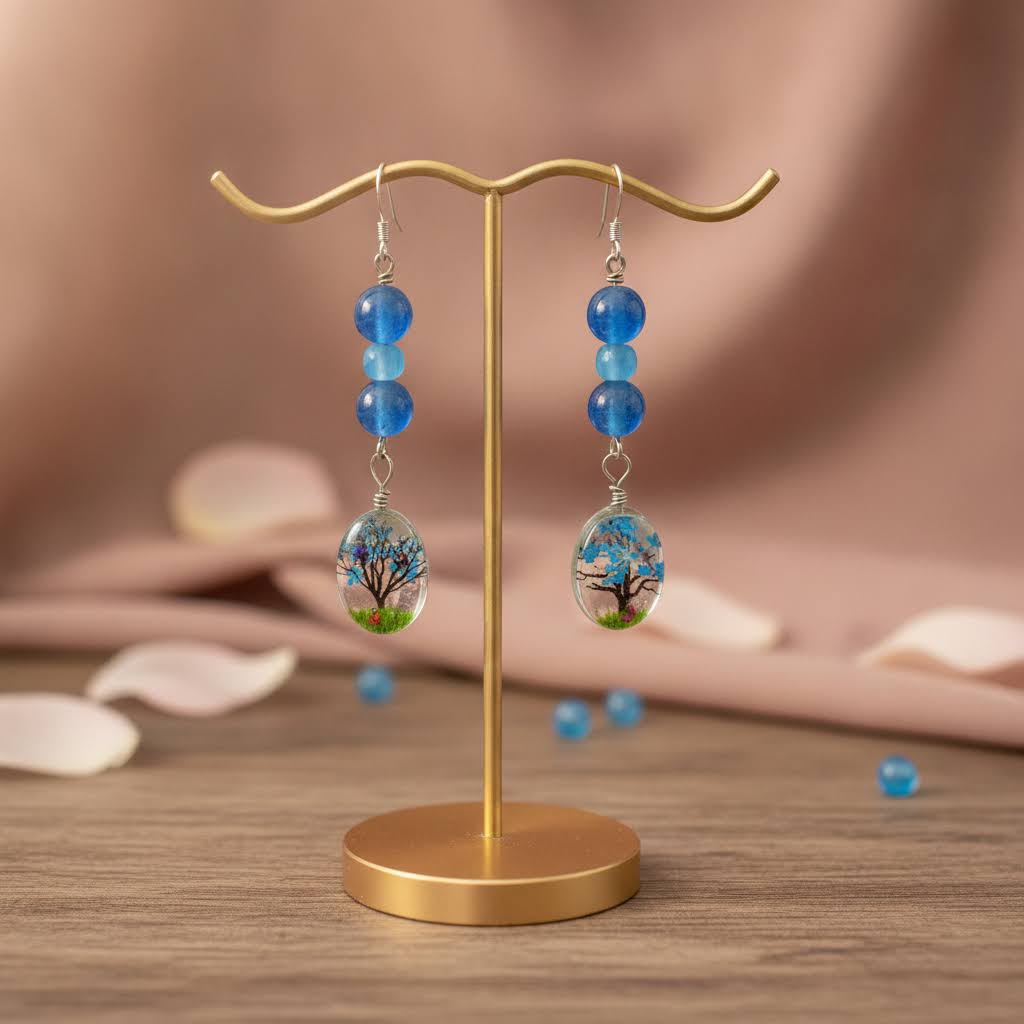 A pair of dangling earrings displayed on a gold stand. Each earring has three blue beads above an oval pendant featuring a colorful tree design with blue sky, green grass, and brown branches.