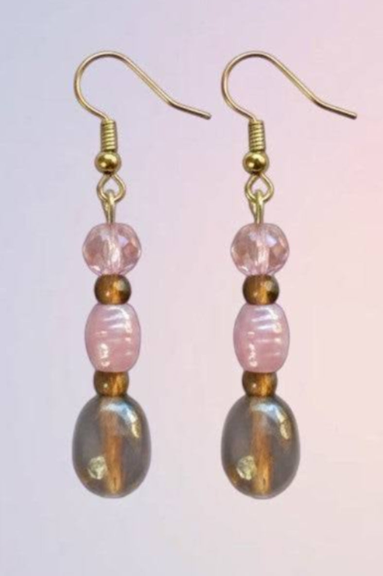 A pair of handmade dangle earrings with gold-tone fishhook wires, pink faceted beads, and teardrop-shaped iridescent gray beads, shown against a soft pink and blue gradient background.