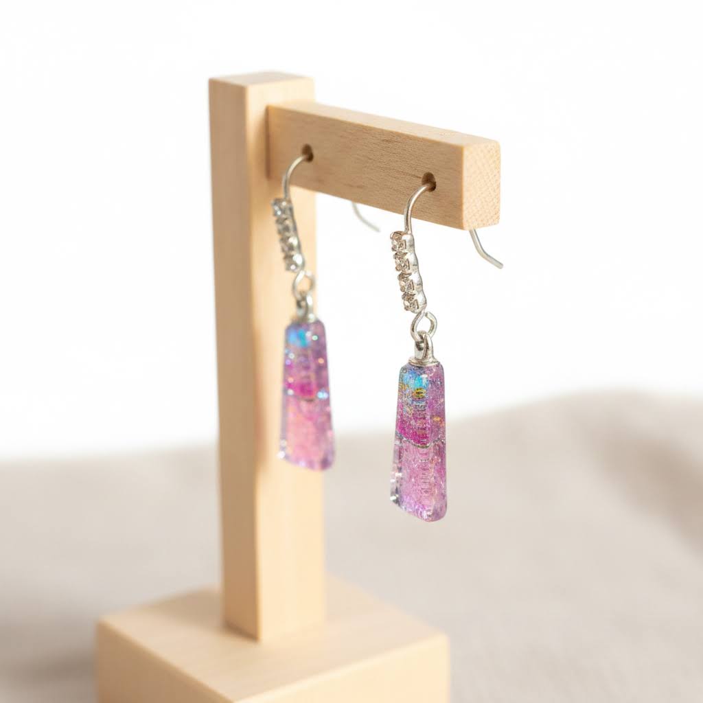 A pair of dangling earrings with rectangular pink and blue glittery charms, displayed on a small wooden stand against a soft white background.