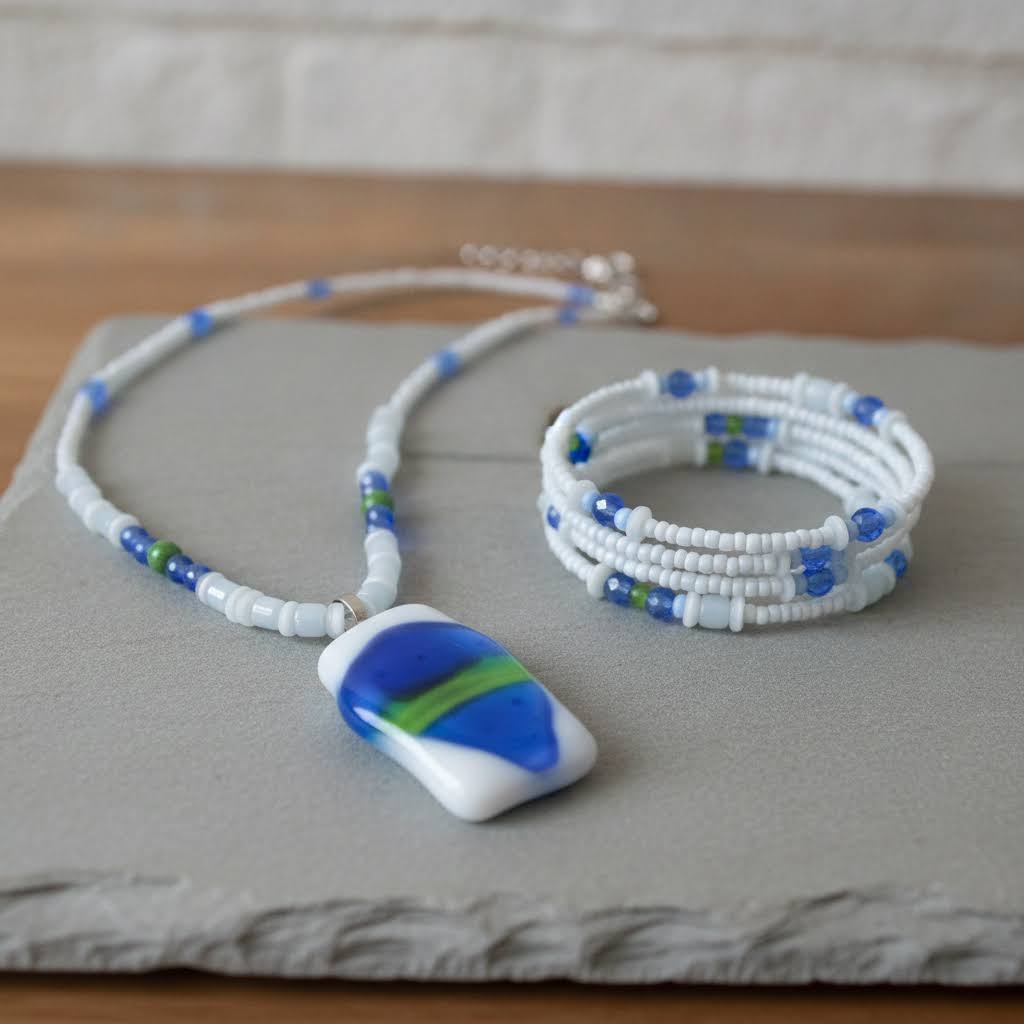 Heartfelt Creations' handmade jewelry set featuring a necklace and a multi-strand bracelet, both crafted with a mix of small, round, white, blue, and green beads, with the necklace featuring a prominent, rectangular, fused glass pendant with a vibrant blue, green, and white abstract design.